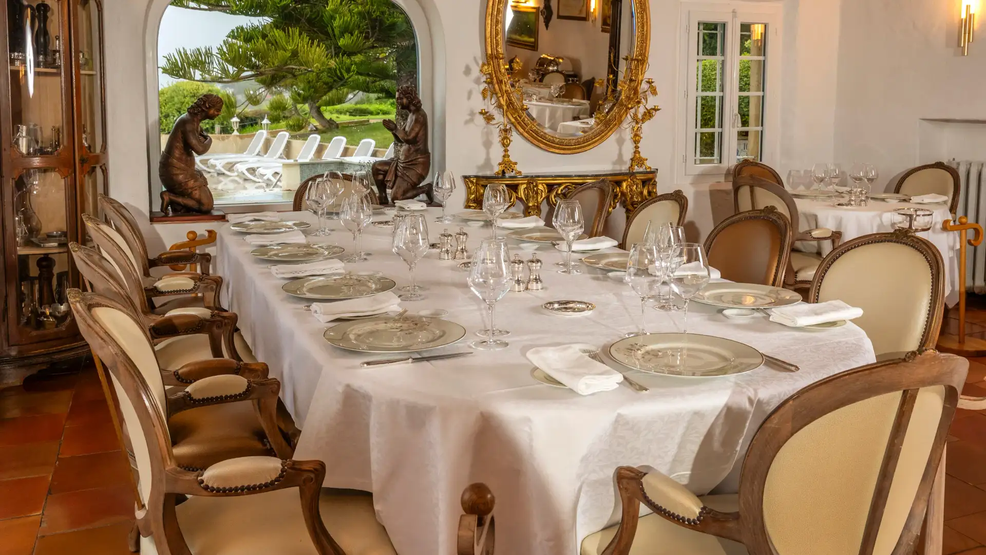 Une table dressée avec des verres et des couverts, entourée de chaises, dans une salle à manger avec vue sur un jardin à travers une grande fenêtre