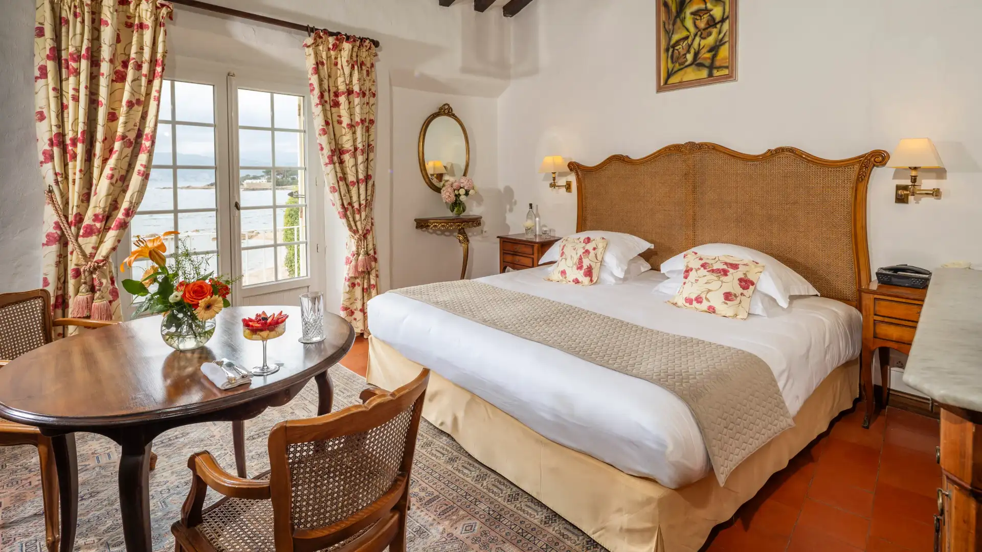 Chambre avec un lit à tête en rotin, une table avec deux chaises et une vue sur la mer à travers une fenêtre aux rideaux floraux