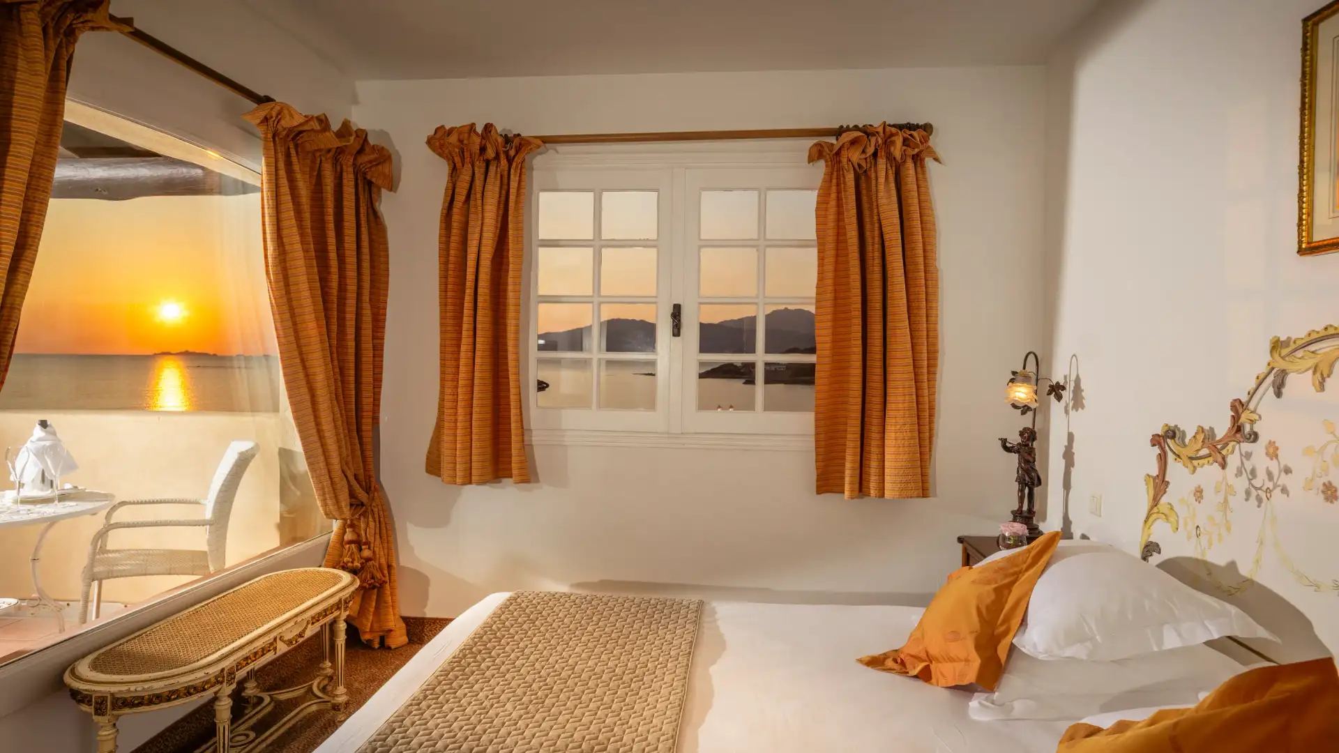 Chambre avec un lit blanc et une couverture beige, une fenêtre avec des rideaux orange et une vue sur un coucher de soleil, une coiffeuse avec un miroir et une chaise, et une lampe de chevet