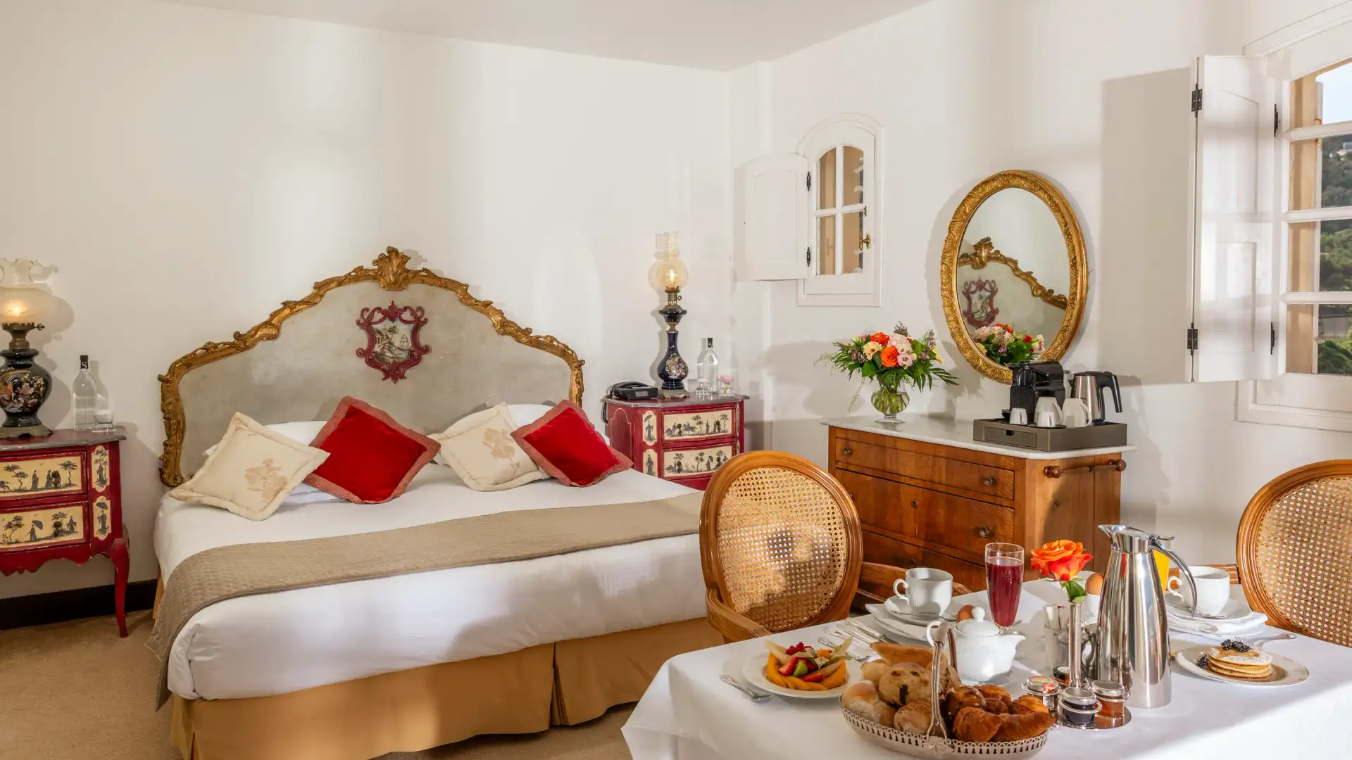 Chambre élégante avec un lit à tête dorée, une table avec un plateau de petit-déjeuner, des meubles anciens et des fenêtres laissant entrer la lumière