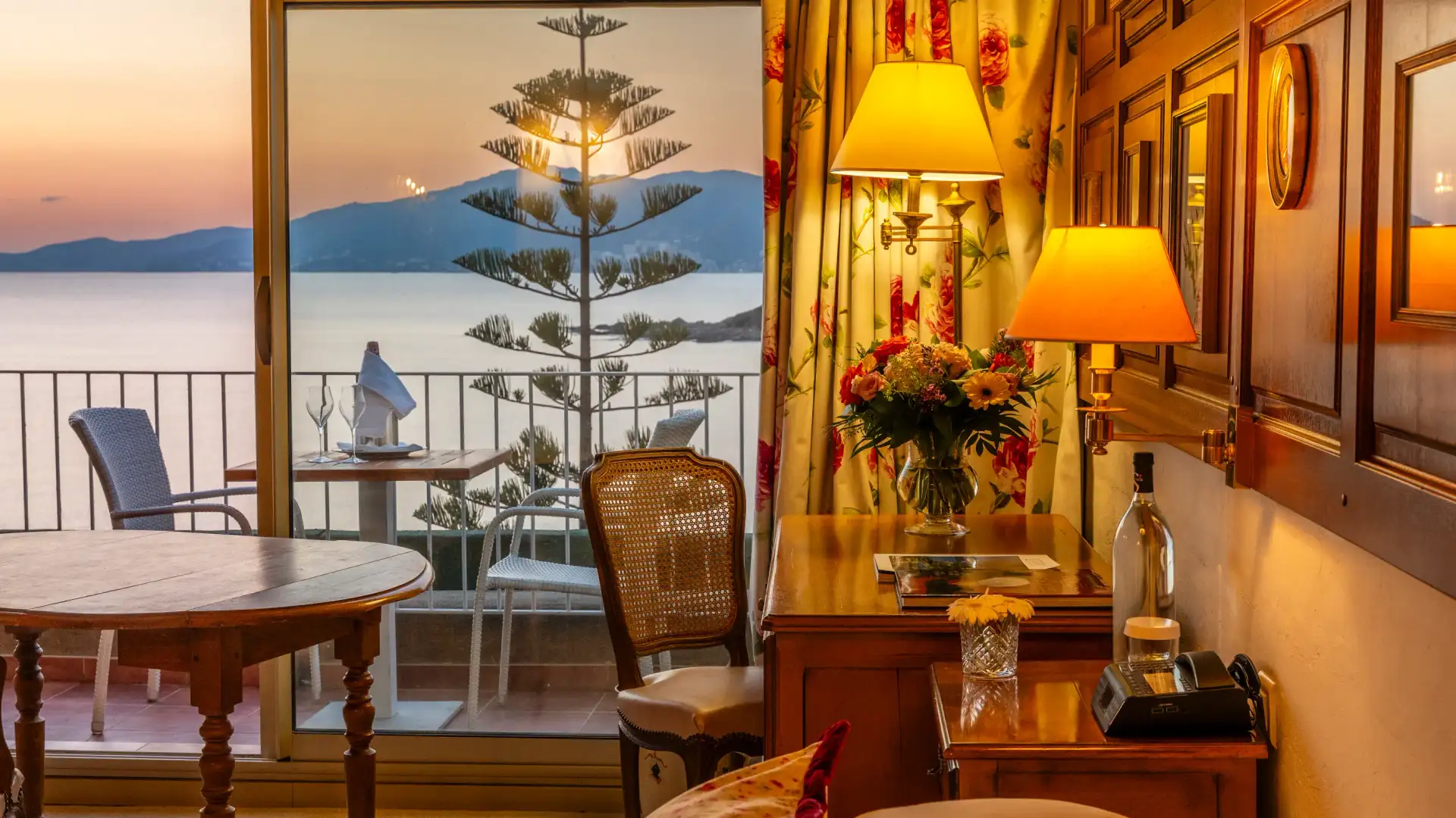 Une pièce avec vue sur mer au coucher du soleil, meublée de tables, chaises et lampes, avec des fleurs sur une commode