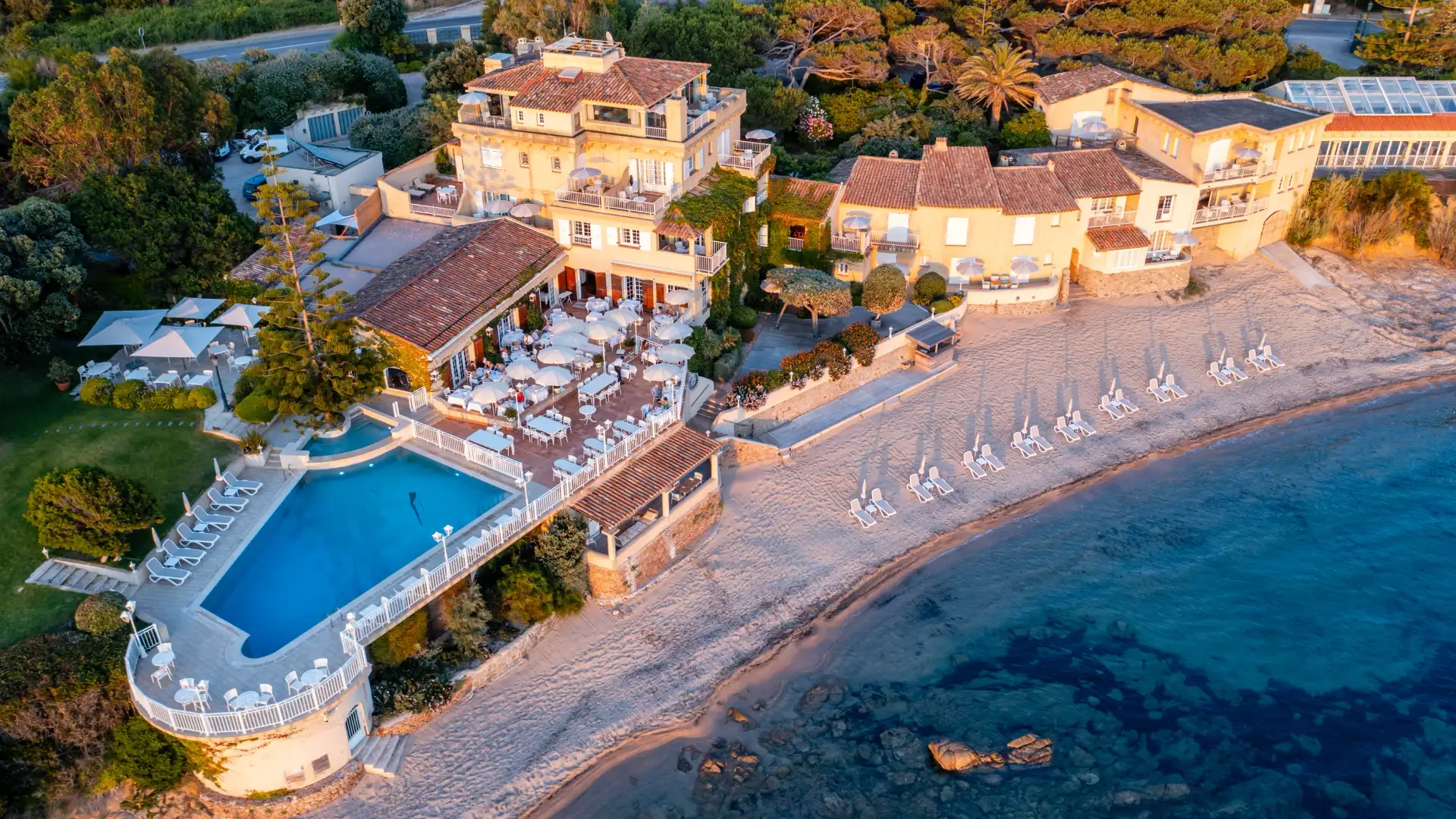 Vue aérienne d'un complexe hôtelier en bord de mer avec piscine, transats et tables en terrasse