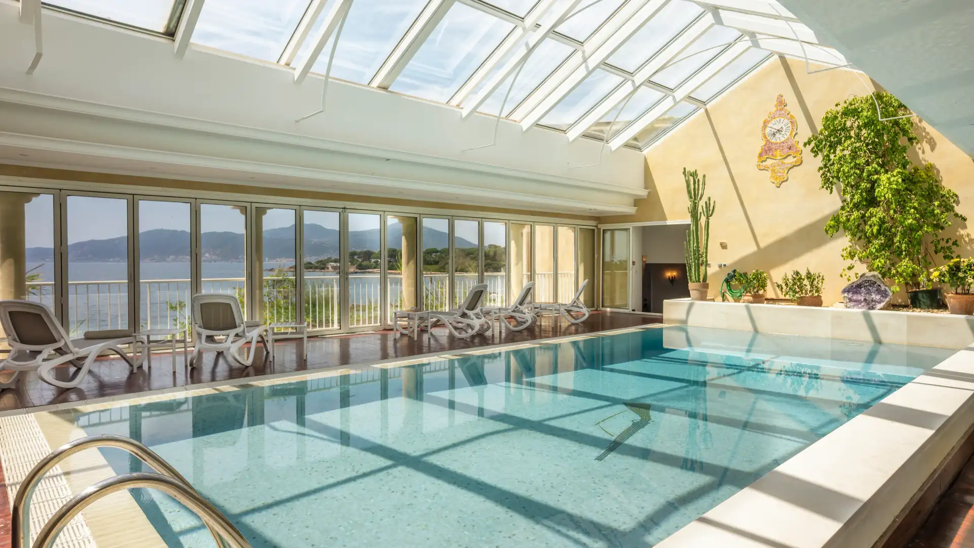 Piscine intérieure avec vue sur la mer et les montagnes, entourée de chaises longues et de plantes décoratives