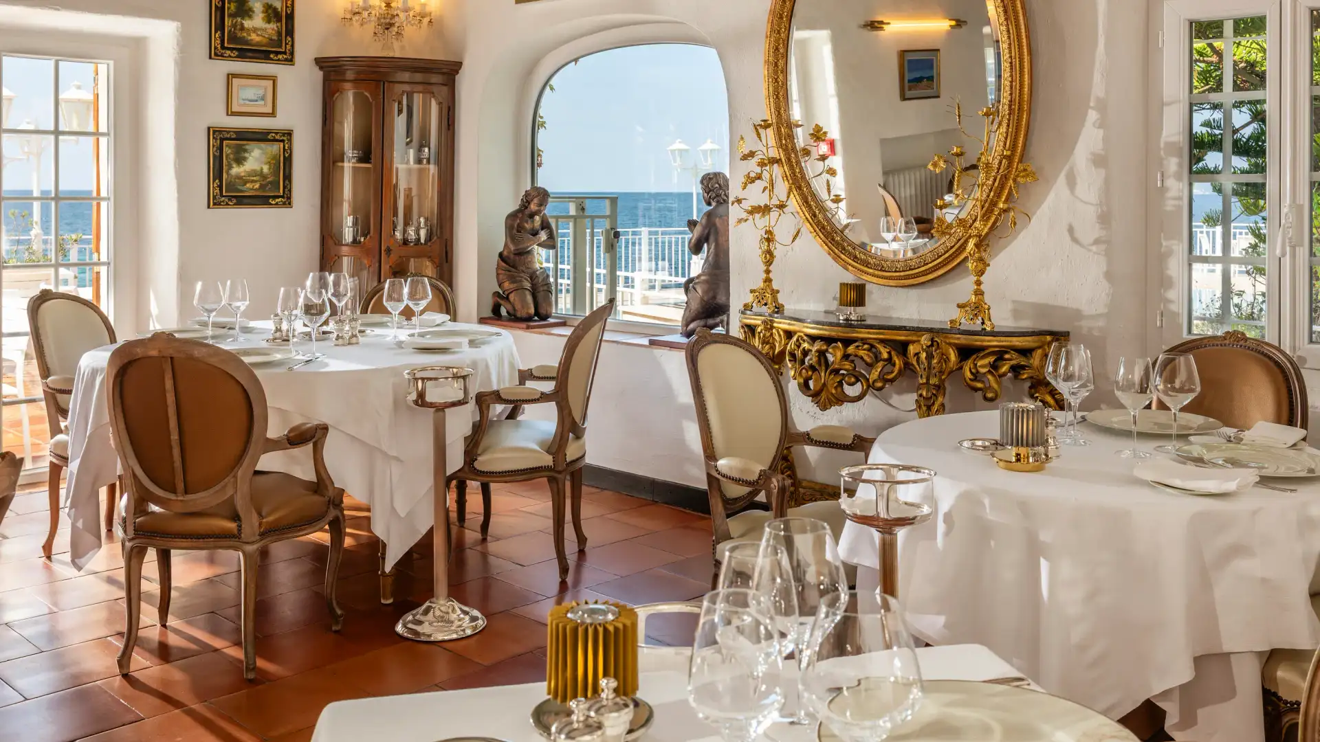 Salle à manger élégante avec tables dressées, chandeliers dorés, statues et vue sur la mer à travers de grandes fenêtres