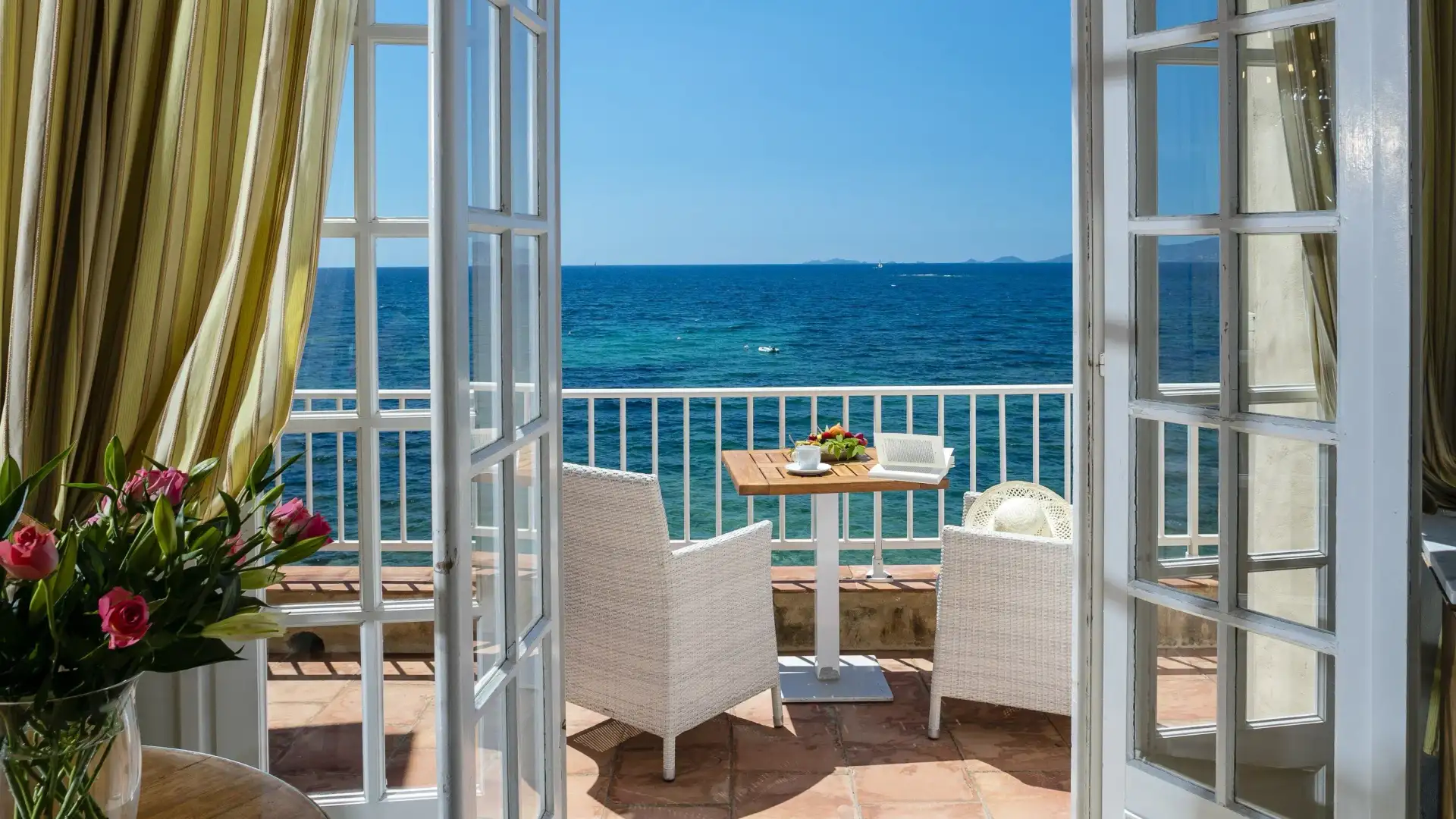 Balcon avec vue sur la mer, table et deux chaises en osier blanc, fleurs roses à gauche, rideaux jaunes à l'intérieur