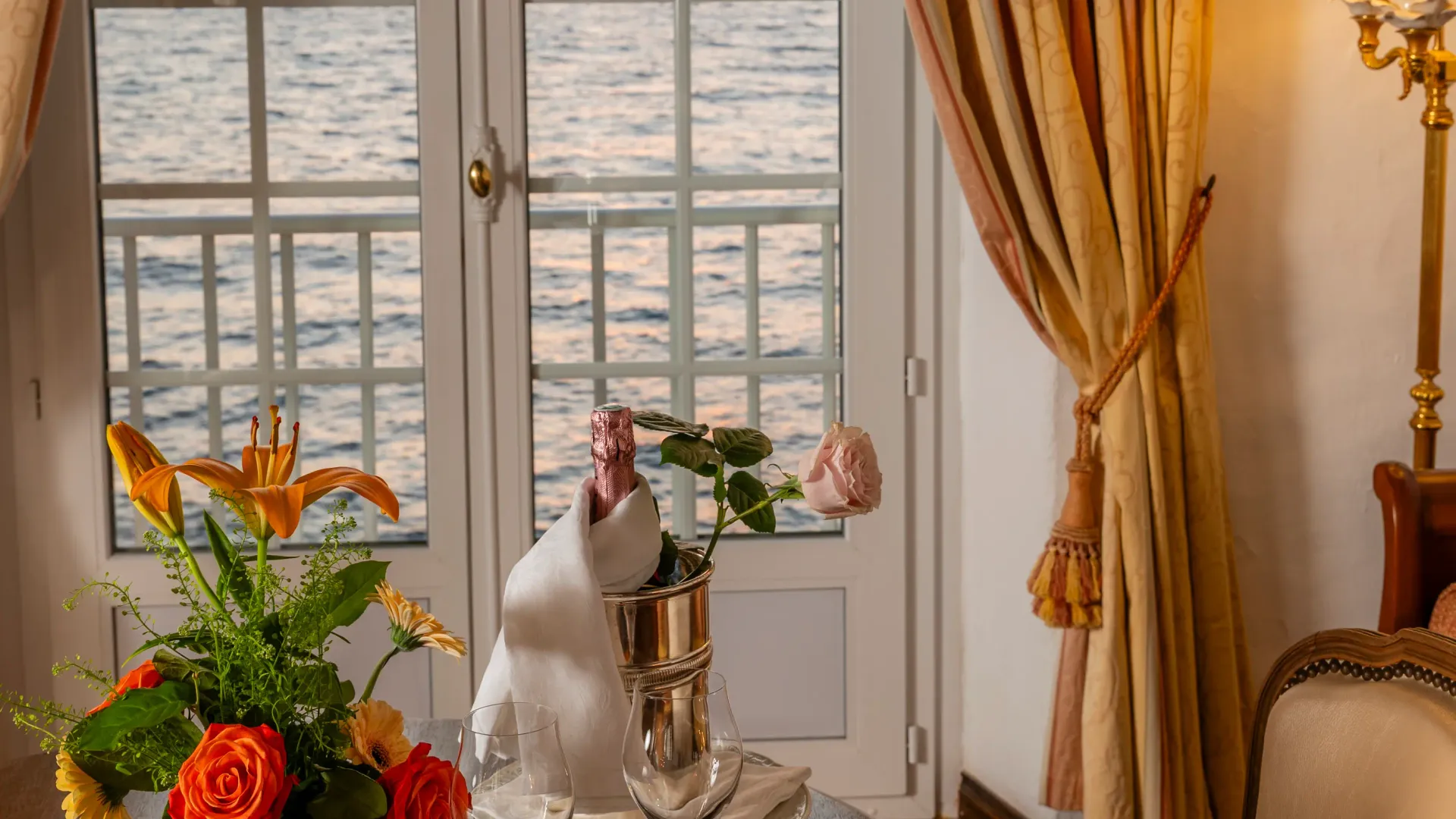 Table avec une bouteille, des verres, une assiette de nourriture, un vase de fleurs et une vue sur la mer au coucher du soleil