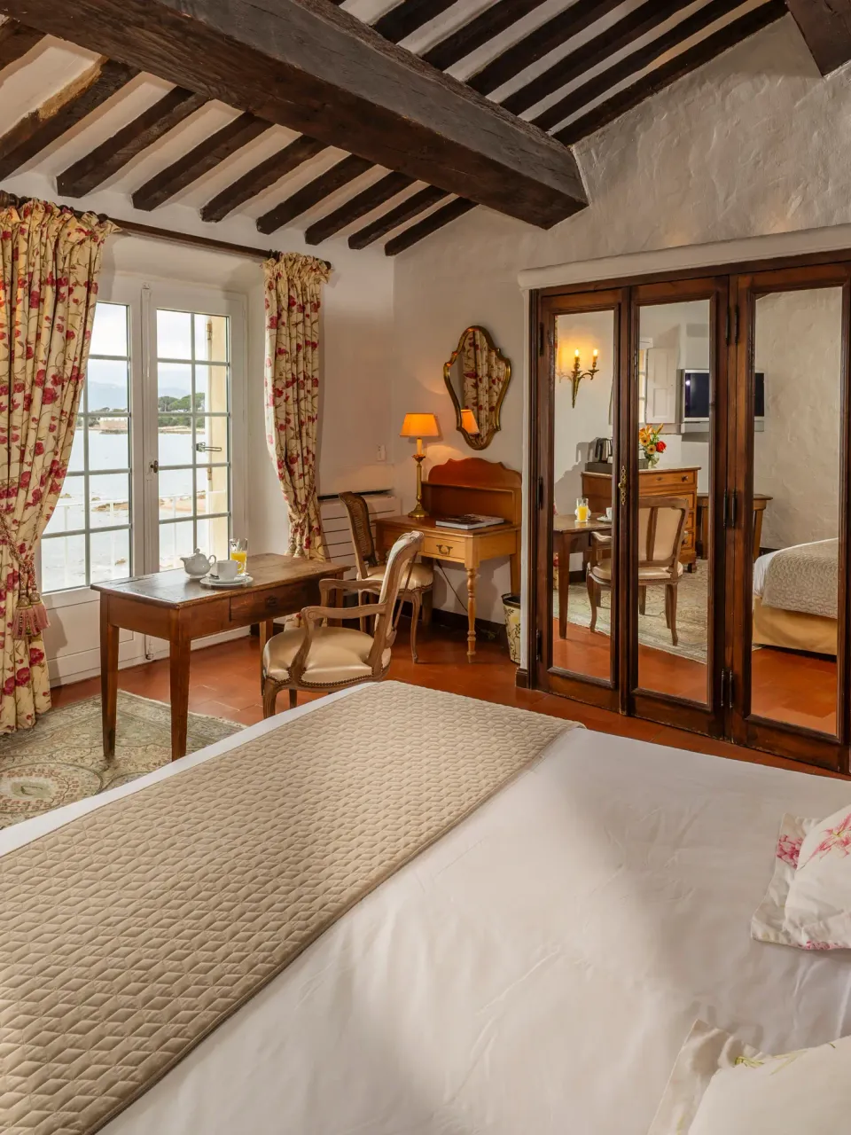 Chambre avec lit double, commode, bureau, miroir et vue sur l'extérieur par les fenêtres
