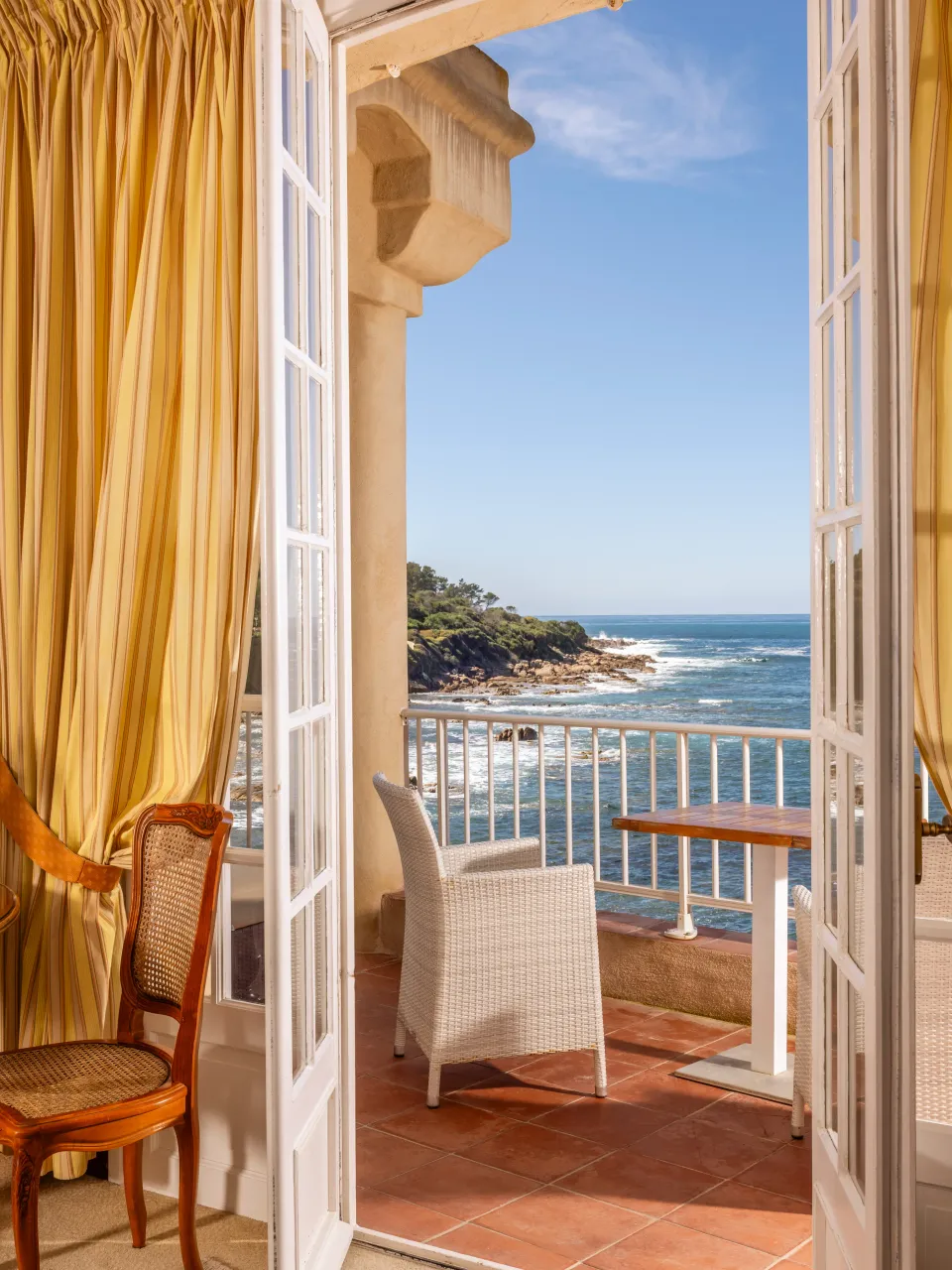Vue depuis une porte-fenêtre ouverte sur une terrasse avec des chaises et une table, donnant sur la mer et des rochers