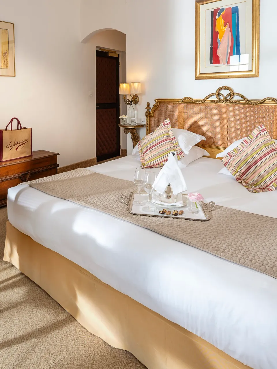 Chambre avec un grand lit à tête en rotin doré, deux chaises en rotin, une table de chevet et des lampes murales, décorée avec des coussins colorés et une serviette pliée