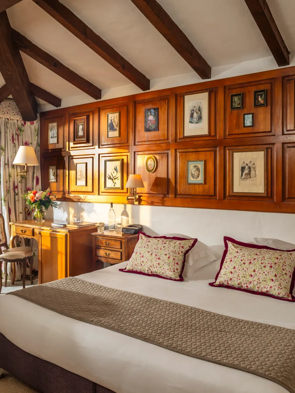 Chambre avec lit double, table et chaises près d'une grande fenêtre offrant une vue sur le paysage extérieur, murs en bois avec cadres et lampes, décoration florale sur les oreillers et la table de chevet