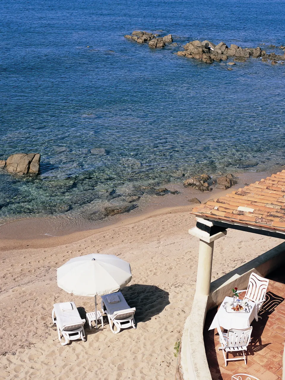 Vue aérienne d'une petite plage de sable avec deux transats sous un parasol et une table dressée sur une terrasse en bord de mer